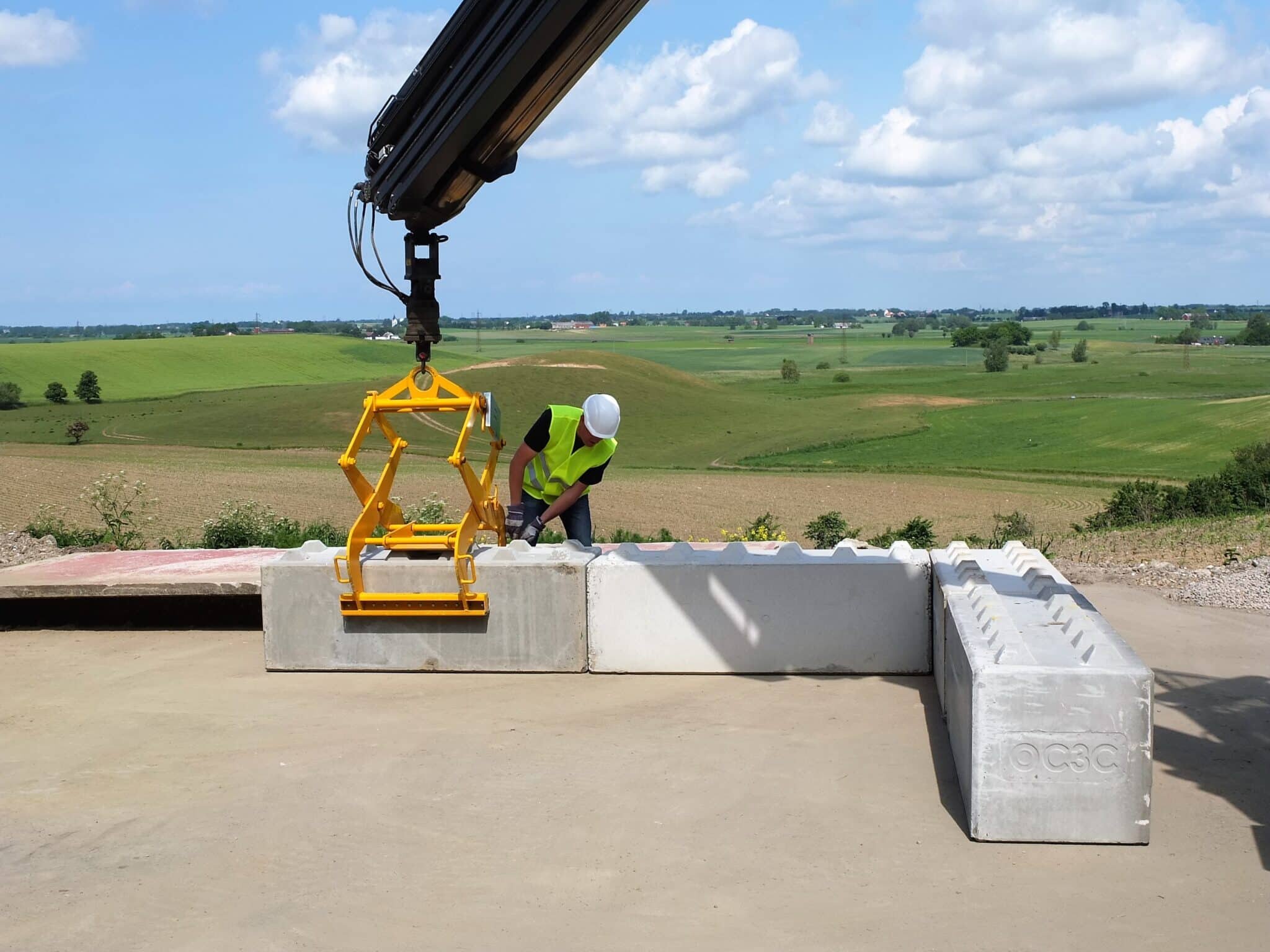 Et bilde av en betongblokker løftetang fra BSV som løfter ett av flere betongelementer. En mann med hjelm er også på bildet og fester løftetangen til betongelementet.